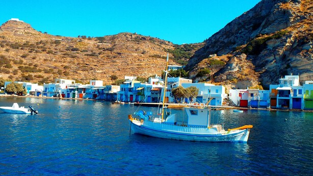 Un poble costaner amb edificis de colors, una embarcació aparcada a l'aigua i un paisatge de muntanyes al fons.