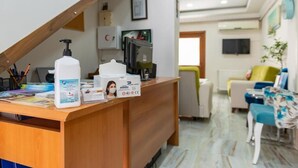 Un mostrador de recepción con un dispensador de gel hidroalcohólico, una caja de pañuelos de papel y una fotografía enmarcada. En el fondo, hay un televisor, una planta y un sofá.