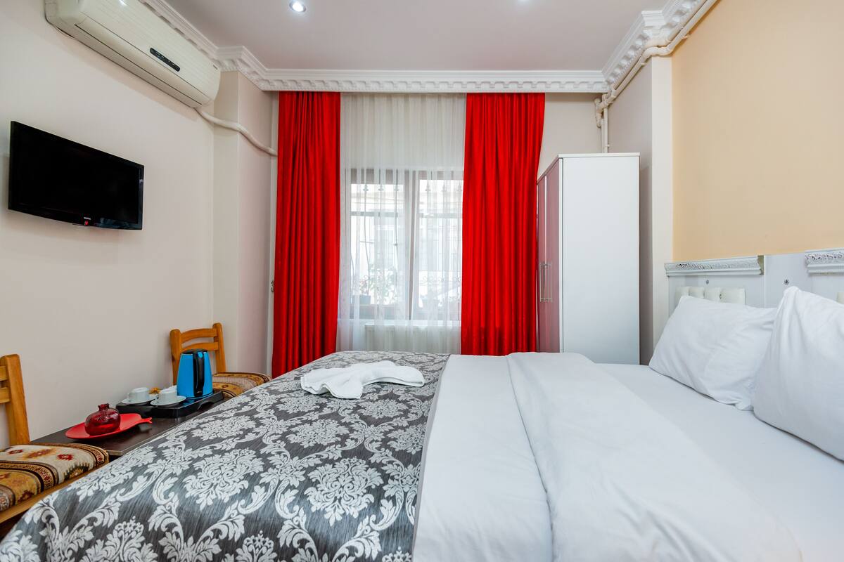 A hotel room with a bed, a TV, a wardrobe, and a window with red curtains.