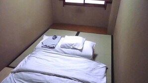 Una habitación pequeña con cama individual y distribución sencilla, incluye una cama con ropa de cama blanca y un almohadón, una mesita con un paño doblado, y una ventana con marco de madera.