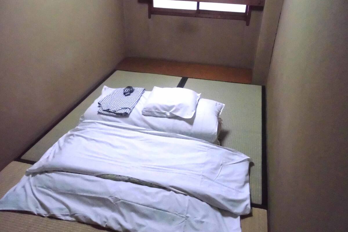 A small, single-bed room with a simple layout, including a bed with white bedding and a pillow, a small table with a folded cloth, and a window with a wooden frame.