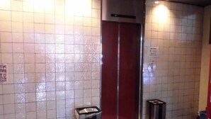 Baño público con pared de azulejos, puerta roja, cubo de basura y un felpudo.