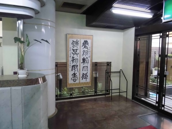 Un área de recepción con mostrador de mármol, un letrero vertical grande con caracteres chinos, una planta en maceta y vistas al exterior con vegetación a través de una puerta de cristal.