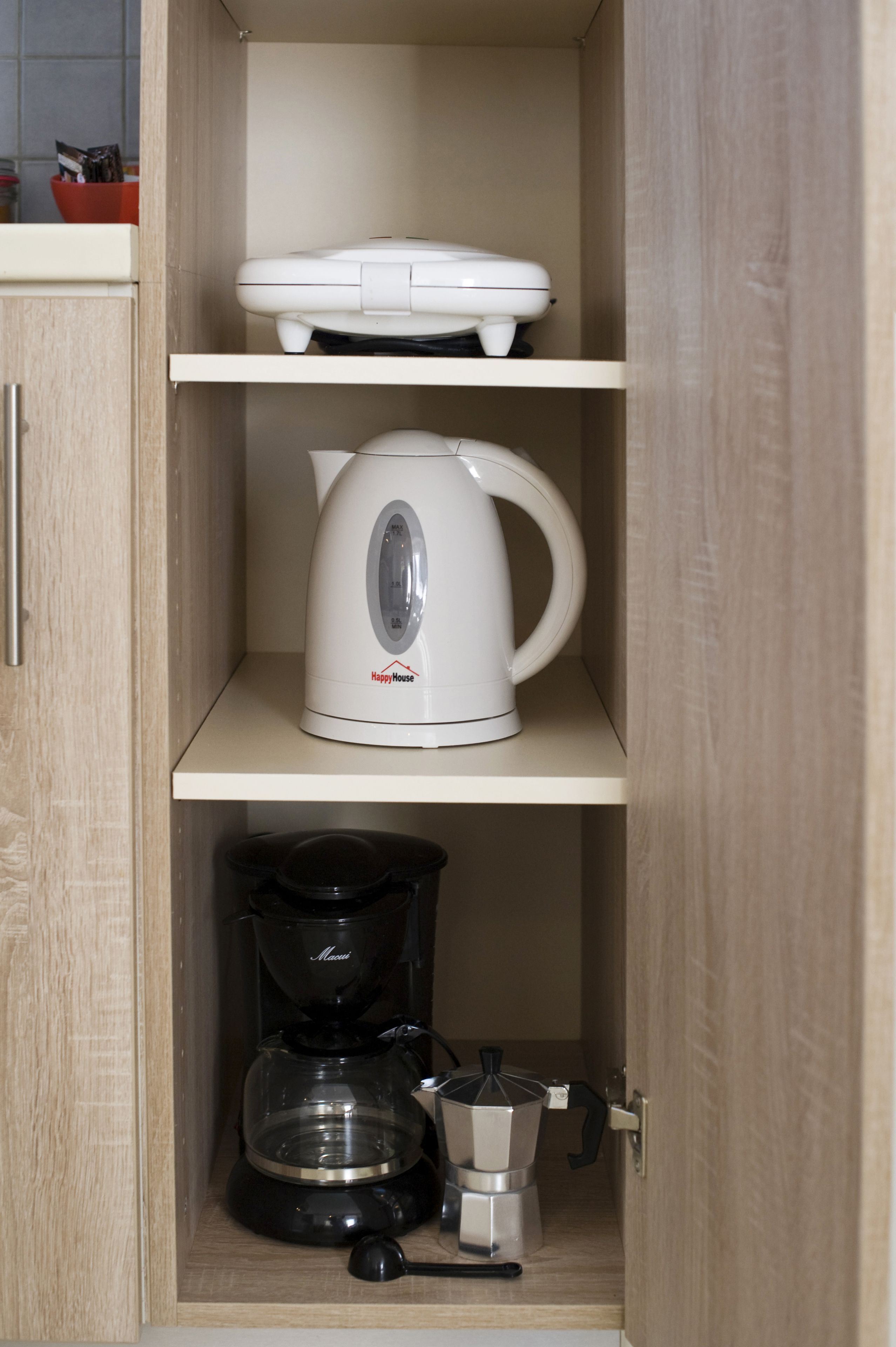 A kitchen cabinet with a white coffee maker on the top shelf, a black coffee maker on the middle shelf, and a coffee pot on the bottom shelf.