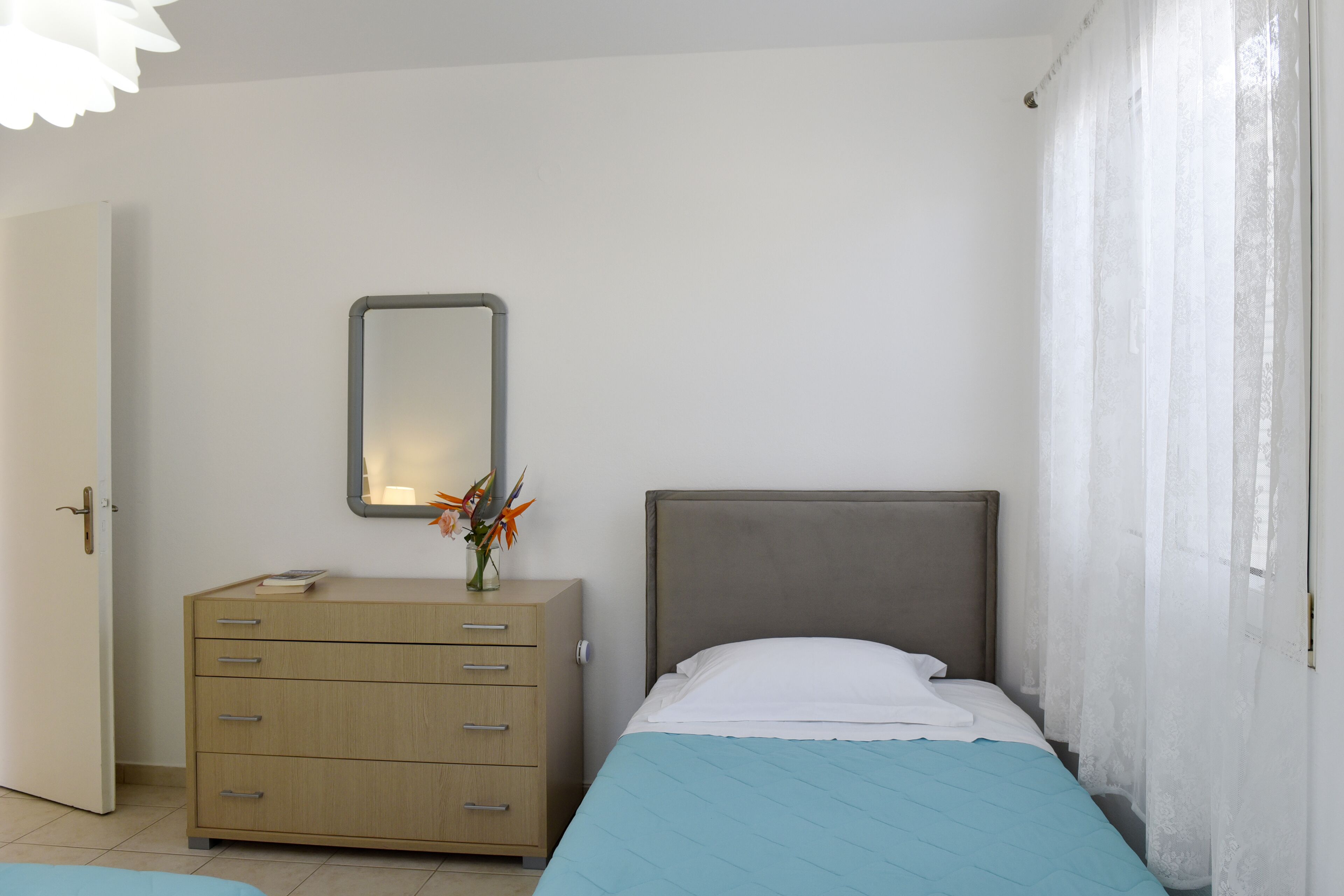 Un dormitorio con cama, cómoda, espejo y ventana con cortinas traslúcidas.
