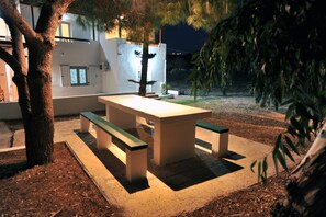 Un área de comedor al aire libre con una mesa y bancos, rodeada de árboles y un edificio al fondo.