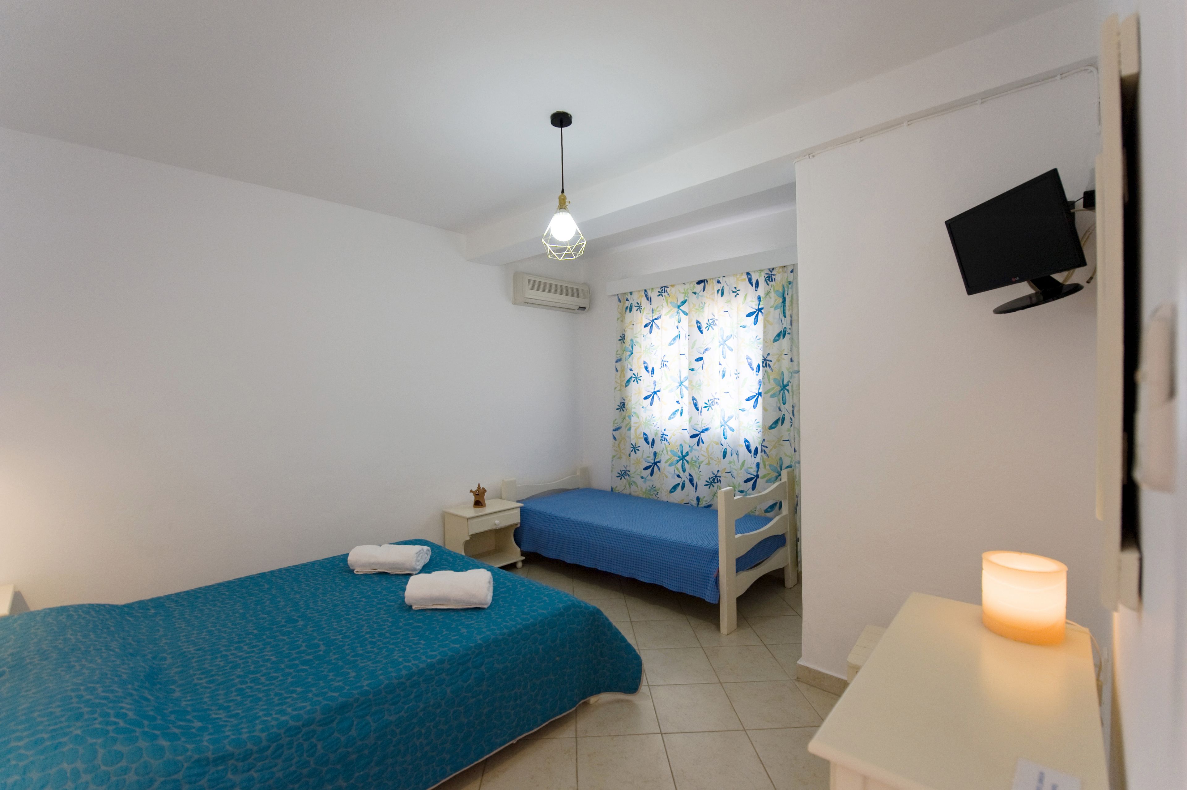 Une petite chambre propre, équipée de deux lits simples, d’un bureau, d’un téléviseur fixé au mur et d’un luminaire au plafond.