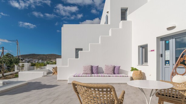A white building with a staircase leading to a balcony. There is a wicker chair and a small table on the balcony. - Tsakanos Home (Milos)