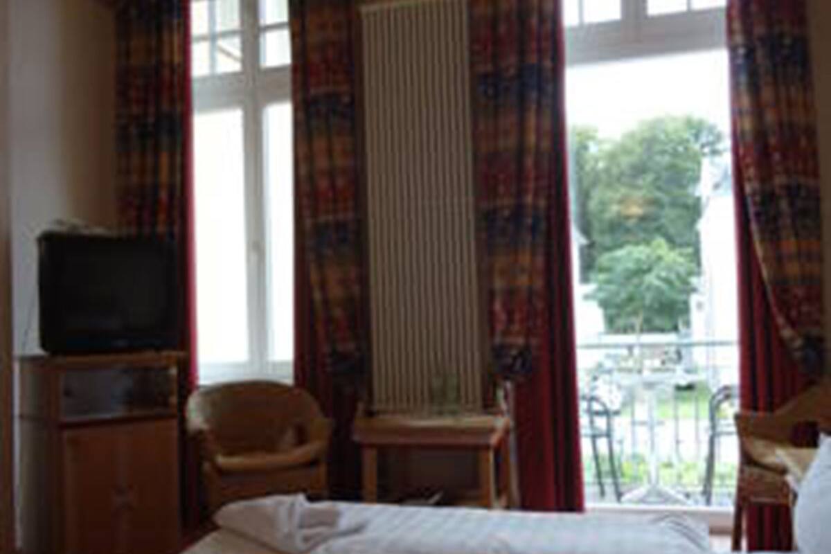 Hotellihuone, jossa on sänky, tuoli, pieni pöytä, televisio ja ikkuna, jossa on verhot.