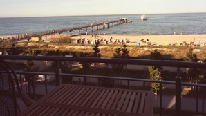 Vistes a una platja amb un moll de fusta que s'endinsa al mar, sorra daurada, persones i gandules, i un cel serè.