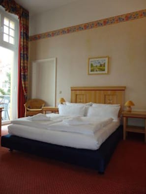 Una habitaci ón de hotel con una cama, una silla, una mesita de noche y un ventanal con cortinas.