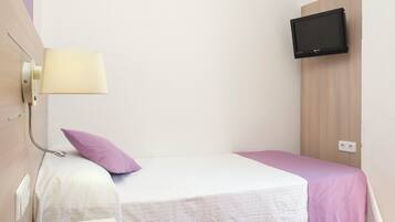 Una cama individual con colcha blanca y morada, una mesita de noche con control remoto, un televisor montado en la pared y una lámpara de pared.