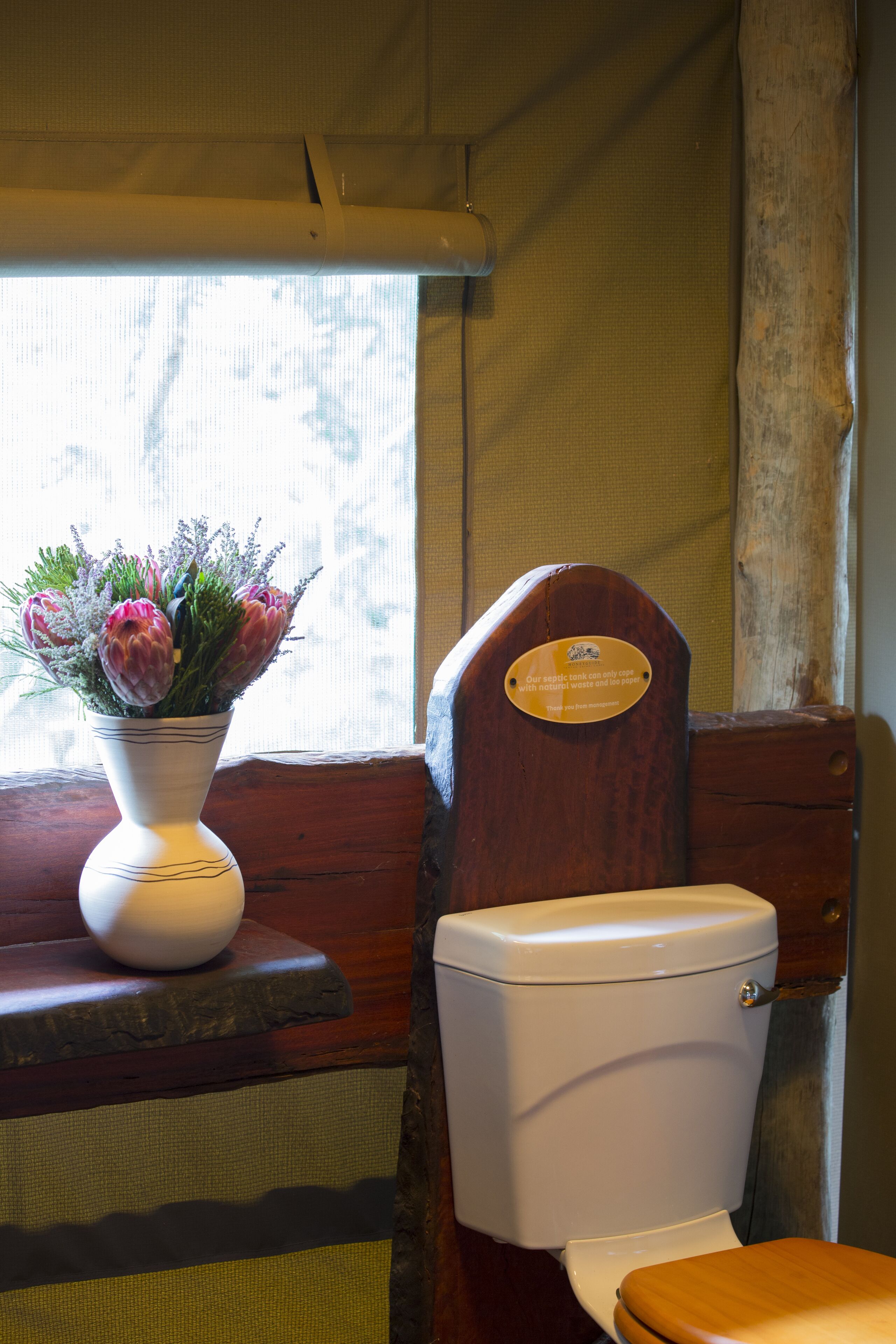 A wooden toilet with a plaque, a white vase with flowers, and a window with a view of trees.
