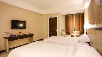 Habitación de hotel con dos camas, un televisor de pantalla plana montado en la pared, una mesita con un jarrón de flores y una puerta que da a otra habitación.