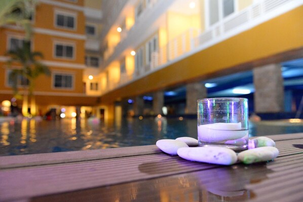 Un vaso de agua sobre una superficie de madera junto a una piscina, con una piedra y un guijarro, con vistas a una zona de piscina y un edificio amarillo al fondo.