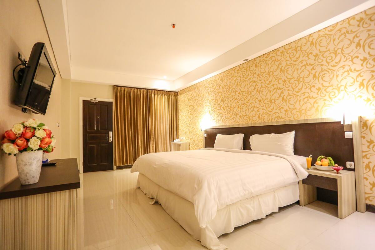 Een hotelkamer met een groot bed, een nachtkastje met fruit, een aan de muur bevestigde televisie en behang met een patroon.