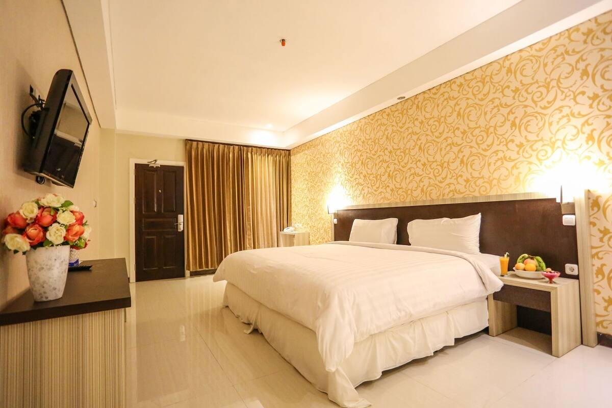 Een hotelkamer met een groot bed, een aan de muur bevestigde televisie, een bloemenvaas en een fruitschaal op het nachtkastje.