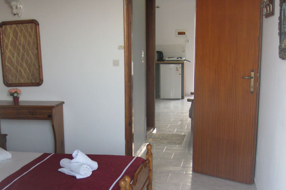 Habitación de hotel con cama, mesita de noche de madera, una pequeña mesa con un jarrón de flores y una puerta que da a una zona de cocina.