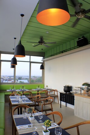 A restaurant with green walls, large windows, and black pendant lights. Tables are set with wine glasses and cutlery.