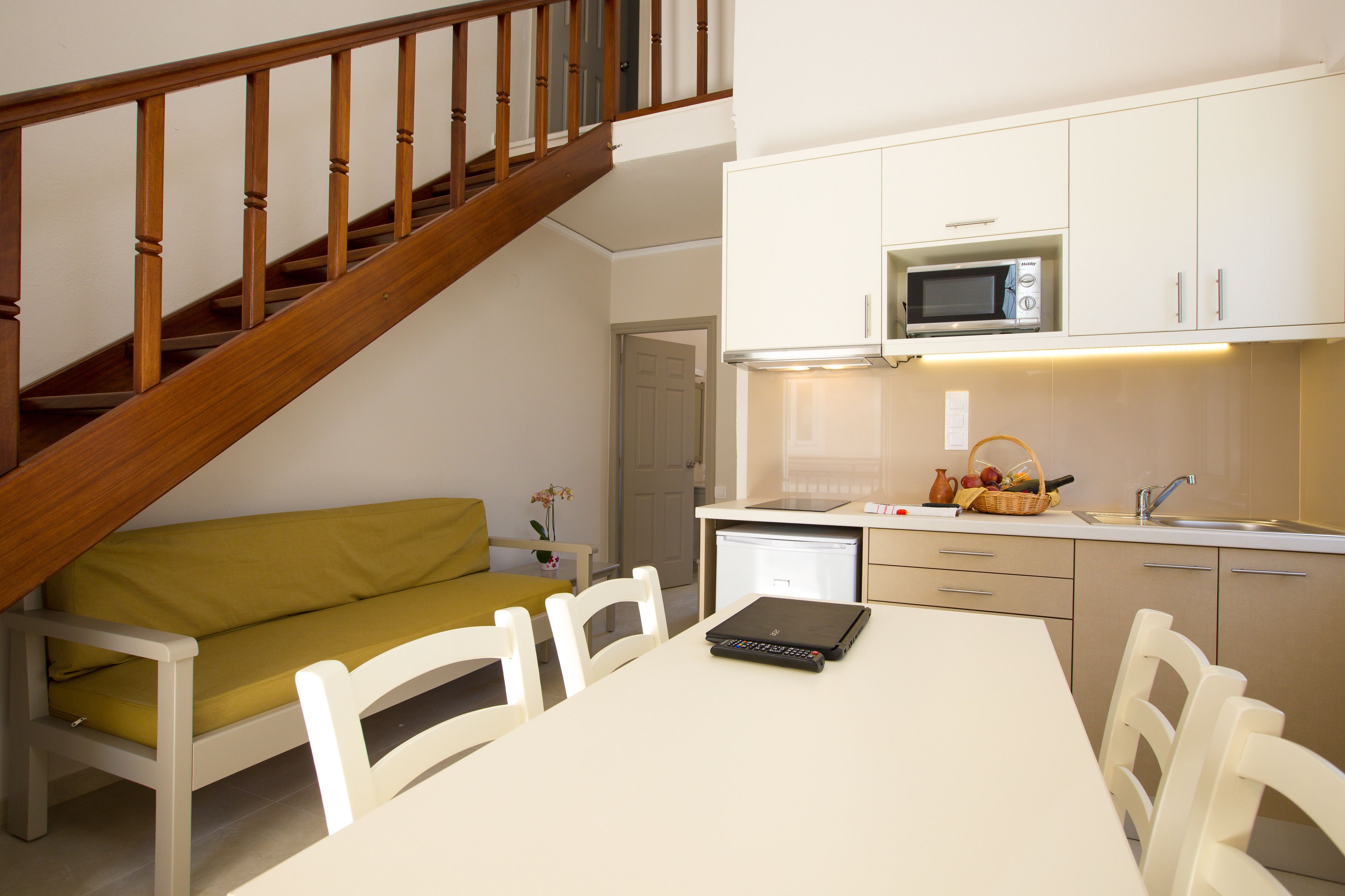 A compact kitchen with white cabinets, a microwave, and a sink. There's a dining table with chairs and a laptop on it. A wooden staircase leads to another room.
