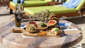 A wooden table with a variety of food items, a bottle of olive oil, and a bottle of water. There are two green lounge chairs and a white umbrella in the background. - Trefon Hotel Apartments and Suites (Rethymno)
