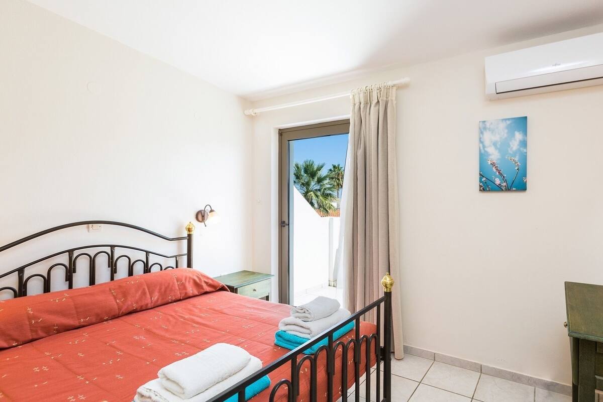 A hotel room with a bed, a desk, an air conditioner, a window with curtains, and a painting on the wall.