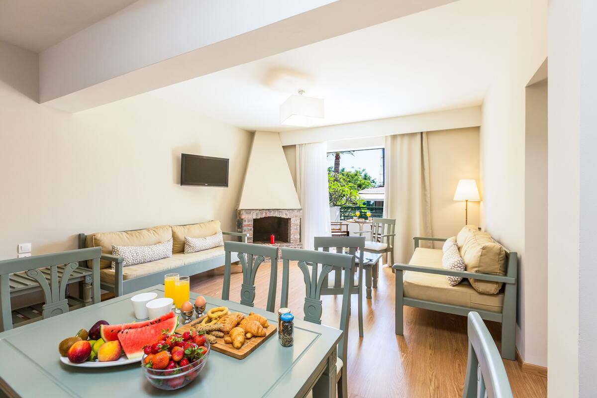 A dining area with a table set for breakfast, including fruits and croissants. There is a sofa and chairs in the background, and a fireplace.