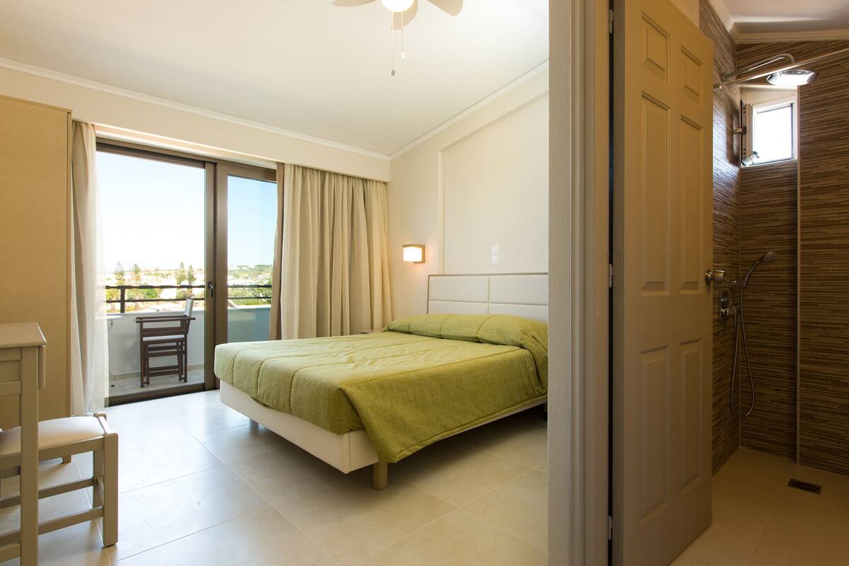 A bedroom with a bed, a chair, a balcony, and a shower area.