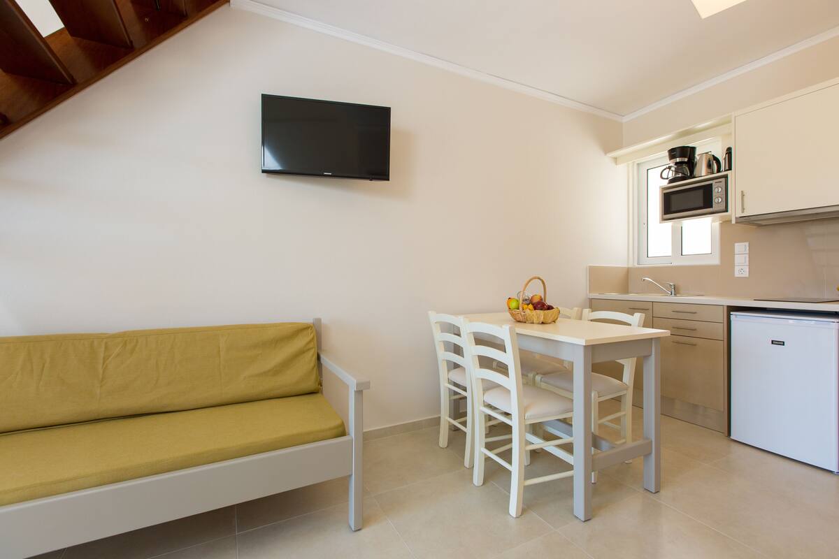 A compact living space with a sofa, a dining table with chairs, a TV mounted on the wall, a microwave, and a small refrigerator.