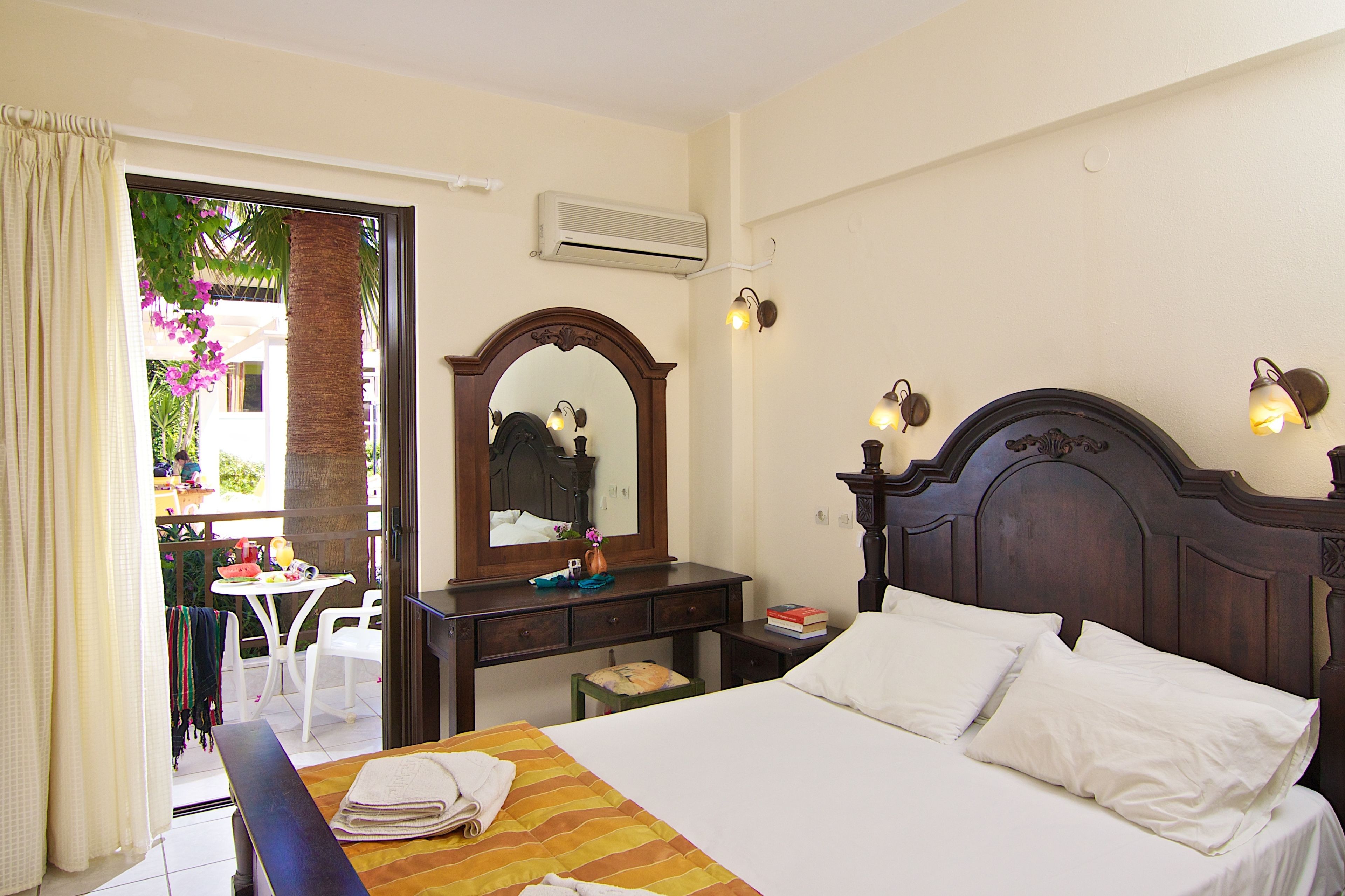 A hotel room with a bed, a wooden headboard, a desk with a mirror, and a view of the outside balcony.