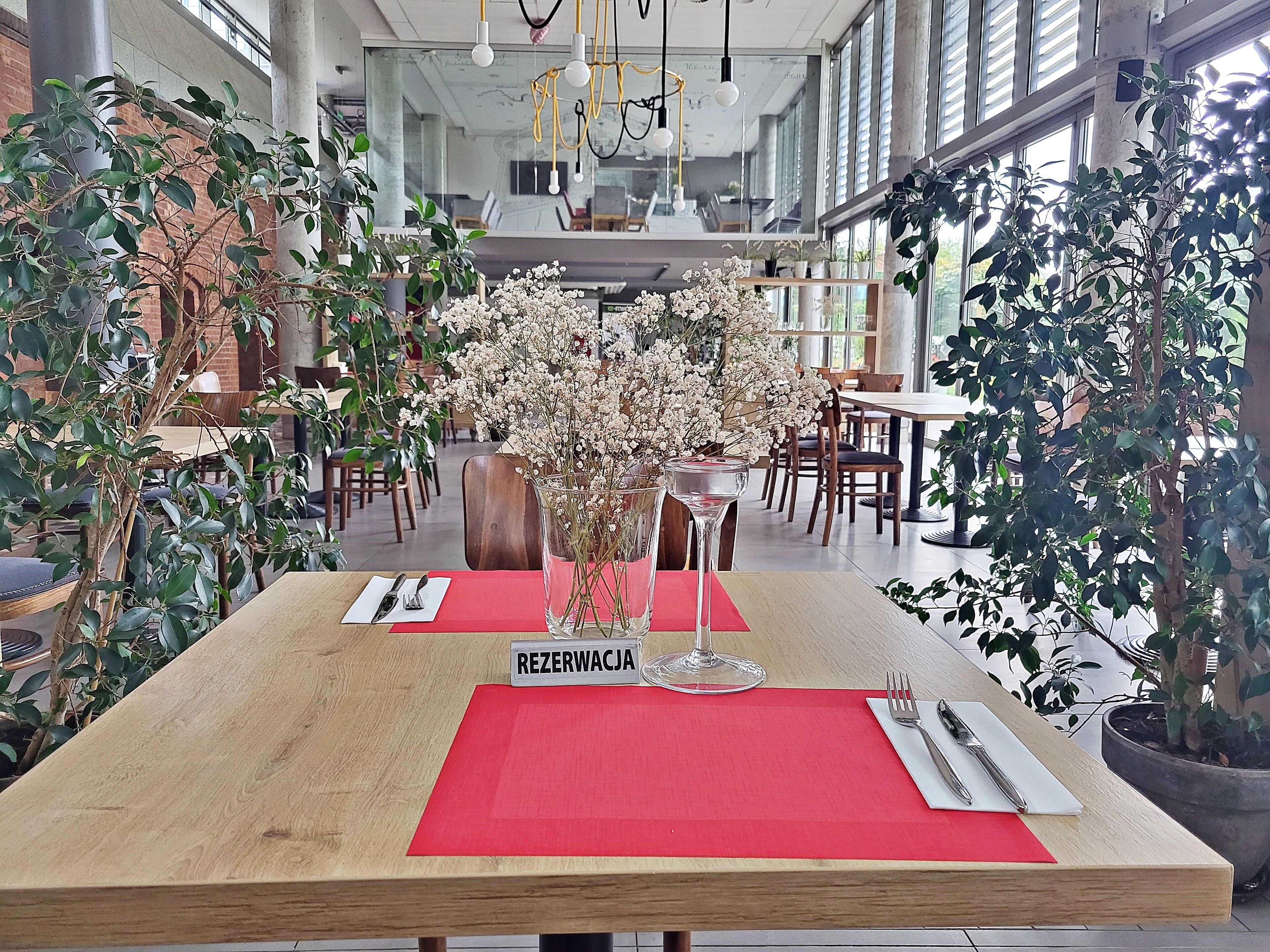 Ein Restaurant mit Holztisch, roten Platzsets, einer Blumenvase und einem Schild mit der Aufschrift 'REZERWACJA'.