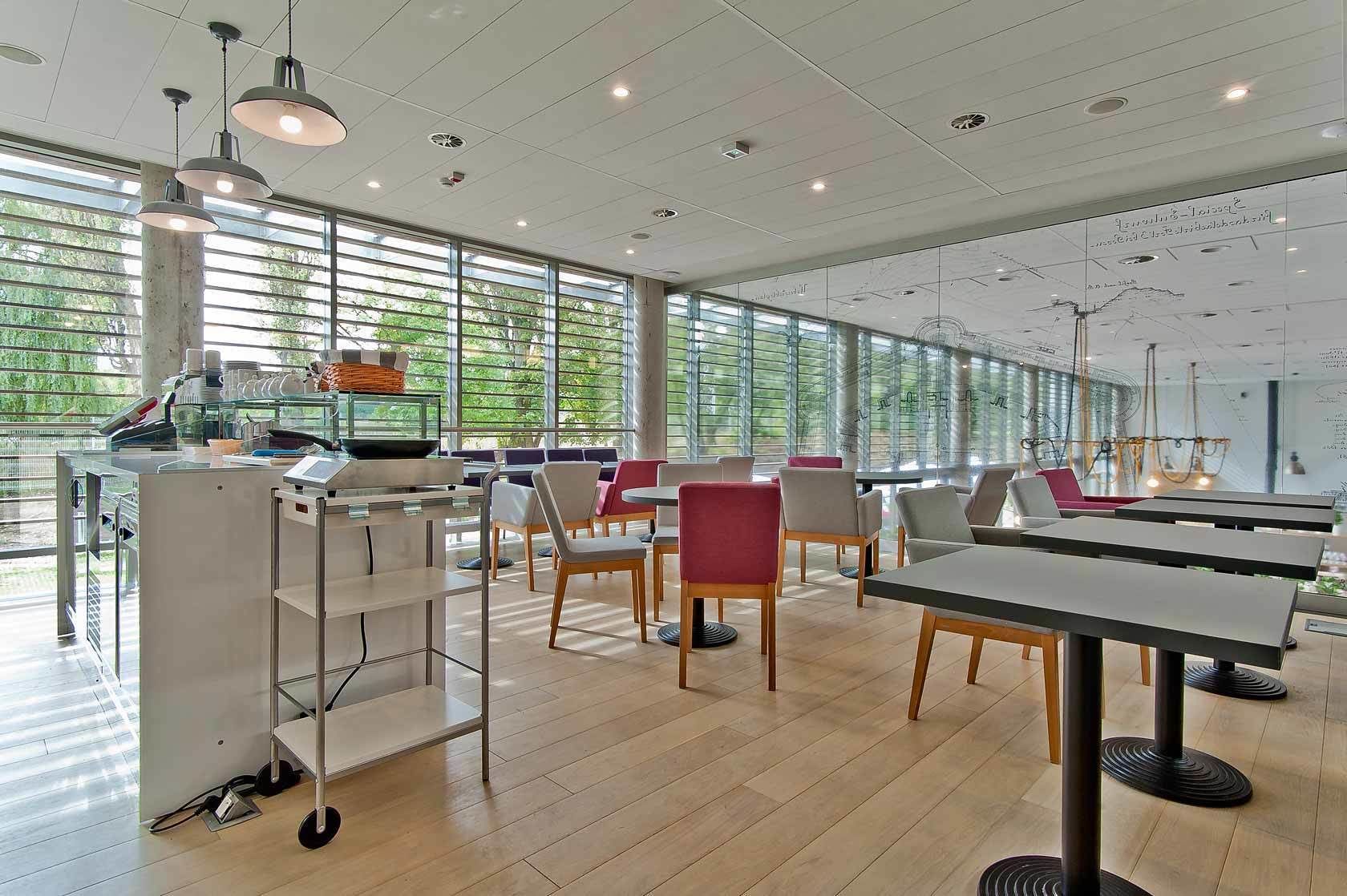 A modern dining area with tables and chairs, large windows, and a counter with kitchen equipment.