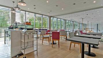 A modern dining area with tables and chairs, large windows, and a counter with kitchen equipment.
