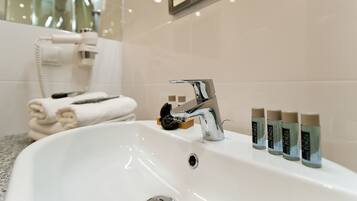 Un baño con un lavamanos blanco, una llave de cromo y un espejo encima. Hay cuatro botellas de loción sobre el lavamanos.
