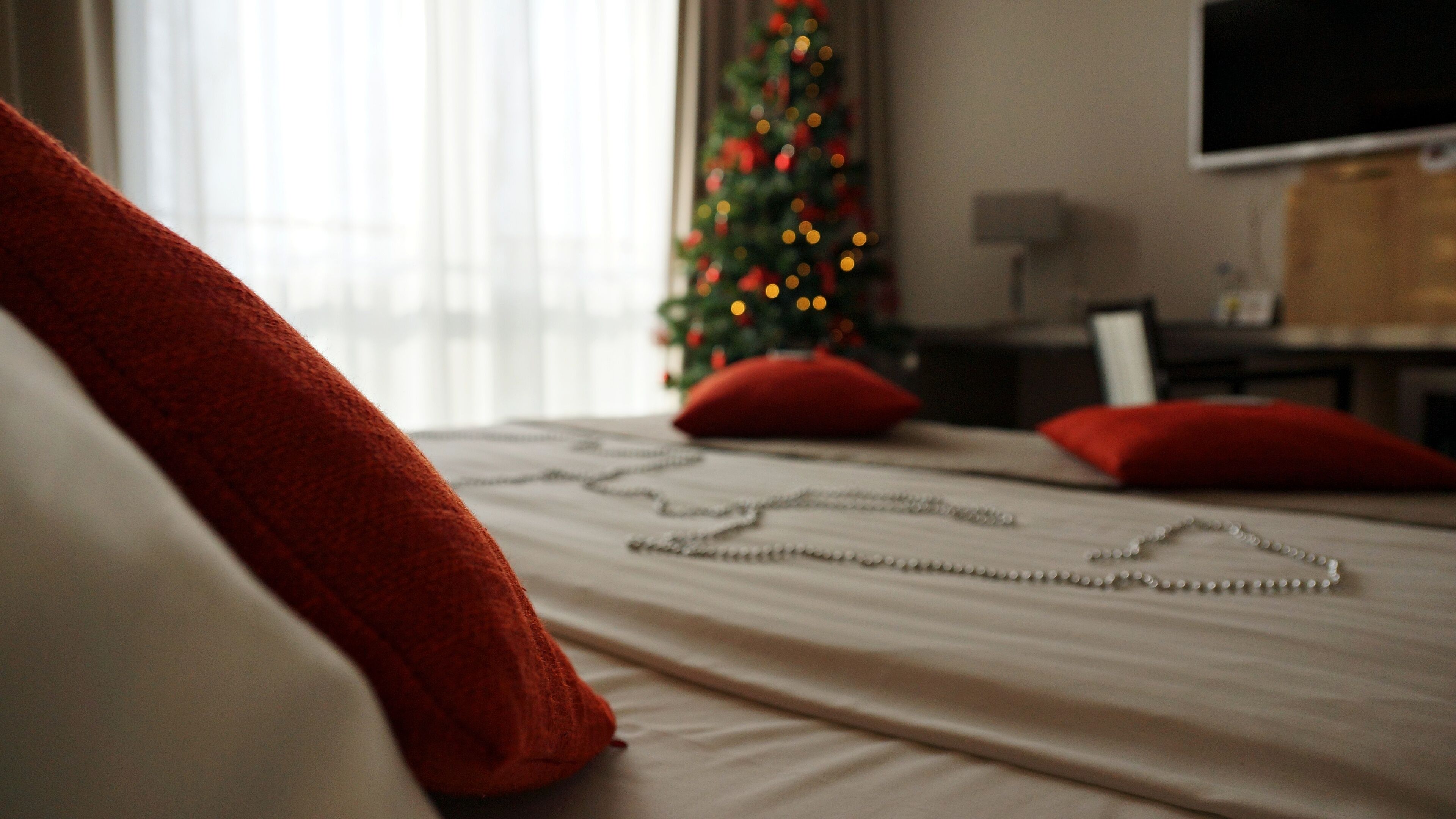 Ein ordentlich gemachtes Bett mit weißer und roter Decke, ein mit Lichtern geschmückter Weihnachtsbaum und ein Fernseher im Hintergrund.