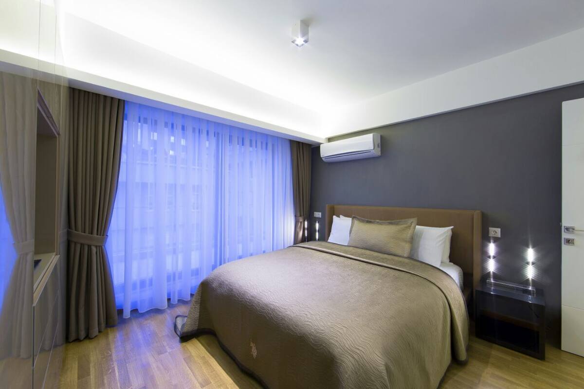 A hotel room with a large bed, a nightstand with lights, an air conditioning unit, and a window with sheer curtains.