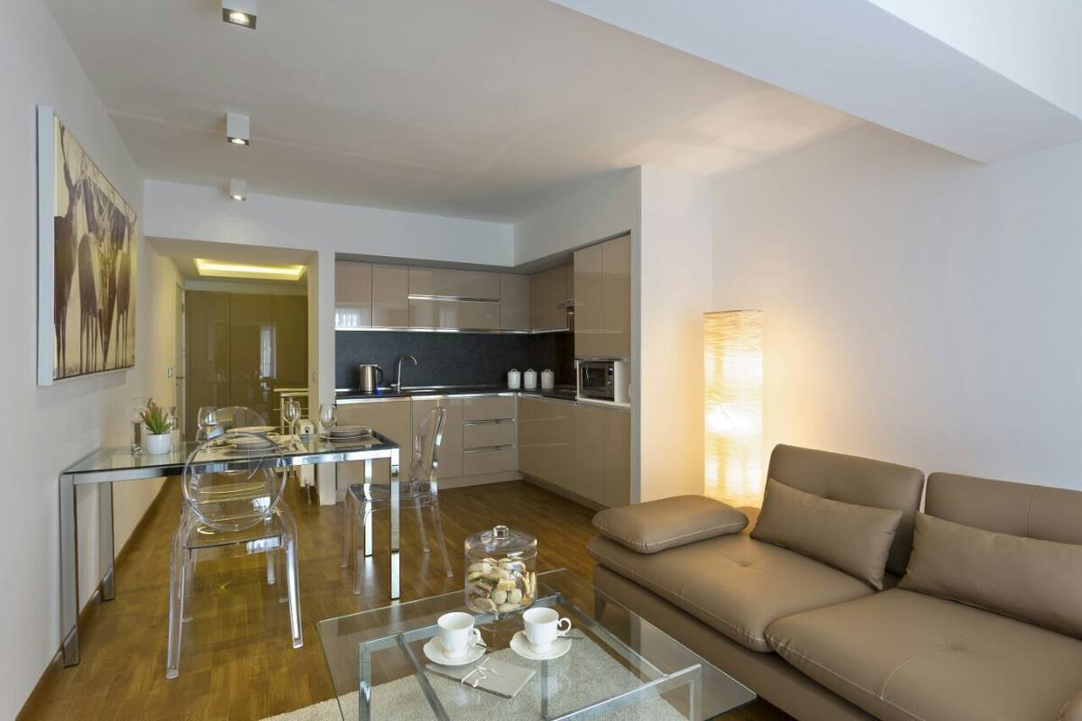 A modern living room with a glass coffee table, a beige sofa, and a kitchen area with stainless steel appliances.
