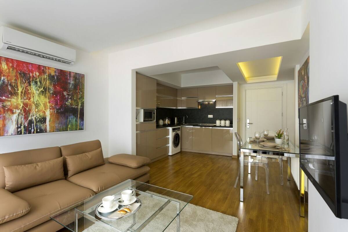 A modern living room with a sofa and a glass coffee table, a kitchen with stainless steel appliances, and a dining area with a table and chairs.