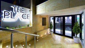 En moderne hotelindgang med et tydeligt skilt, der siger "THE PLACE", trægulv og potteplanter.