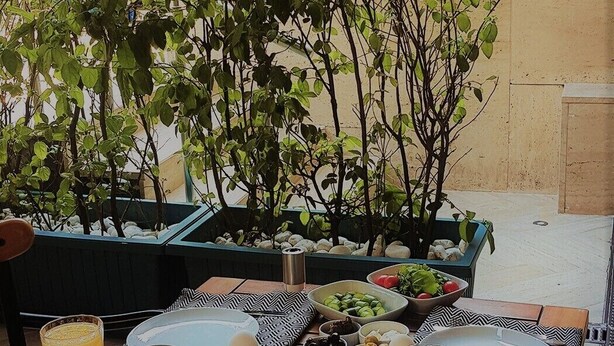 Et udendørs spiseområde med et bord dækket til morgenmad, inklusive æg, ristet brød og frugt. Der er krukker med planter og et skilt i baggrunden, der siger "THE PLACE".