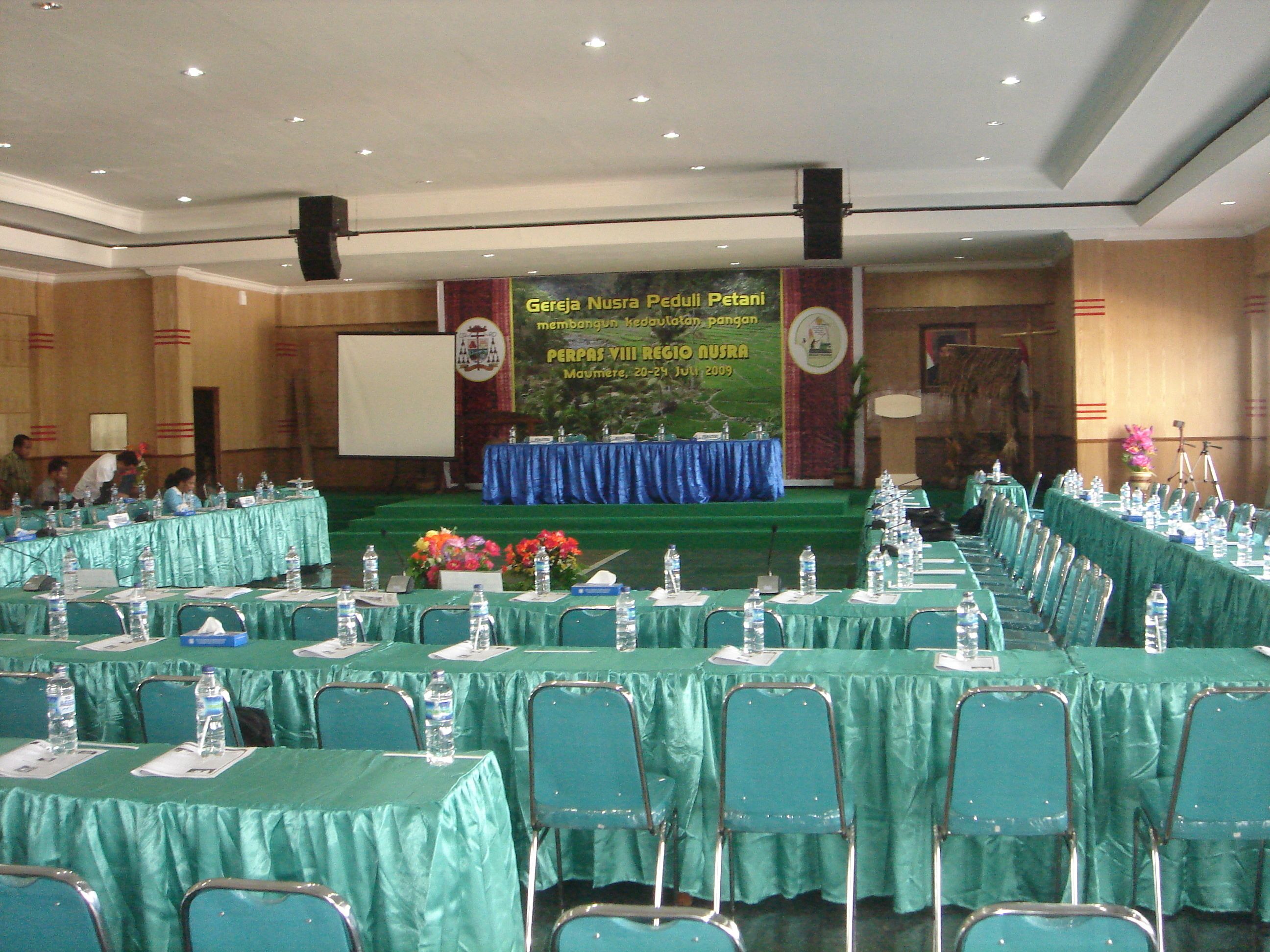 A conference room with rows of tables and chairs, a stage with a banner, and a projector screen.