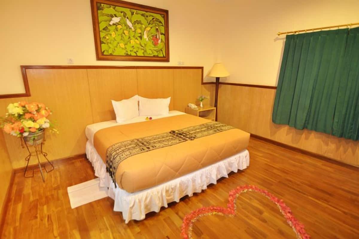A hotel room with a wooden headboard, a bed with white and beige bedding, a bedside table with a lamp, a green curtain, and a floral arrangement on the floor.