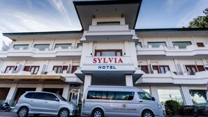 Een hotel genaamd "SYLVIA HOTEL" met een moderne gevel, een overdekte ingang en een parkeerplaats met voertuigen.