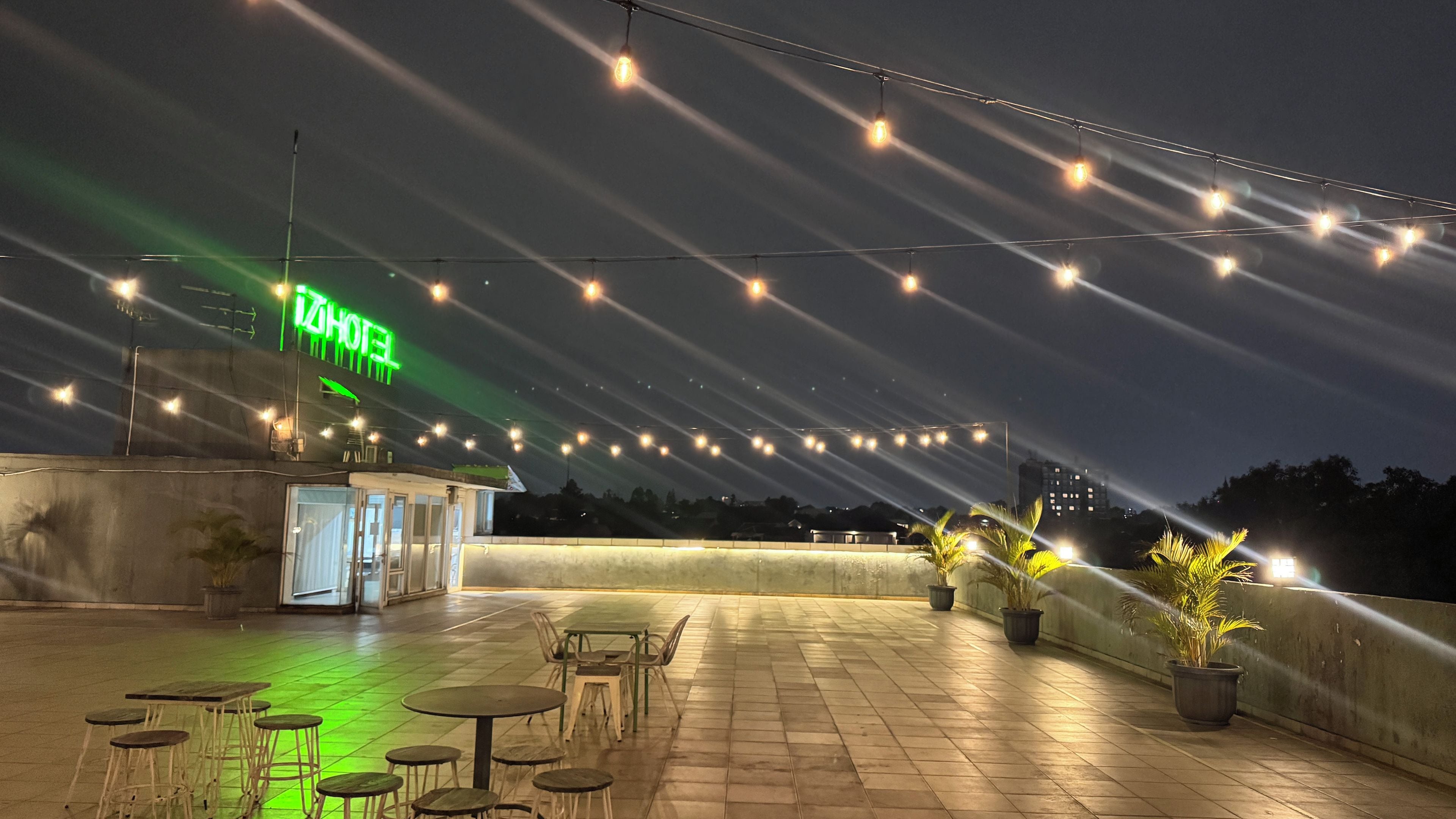 Un espacio al aire libre con mesas y sillas, luces colgantes y un letrero de neón que indica un hotel.