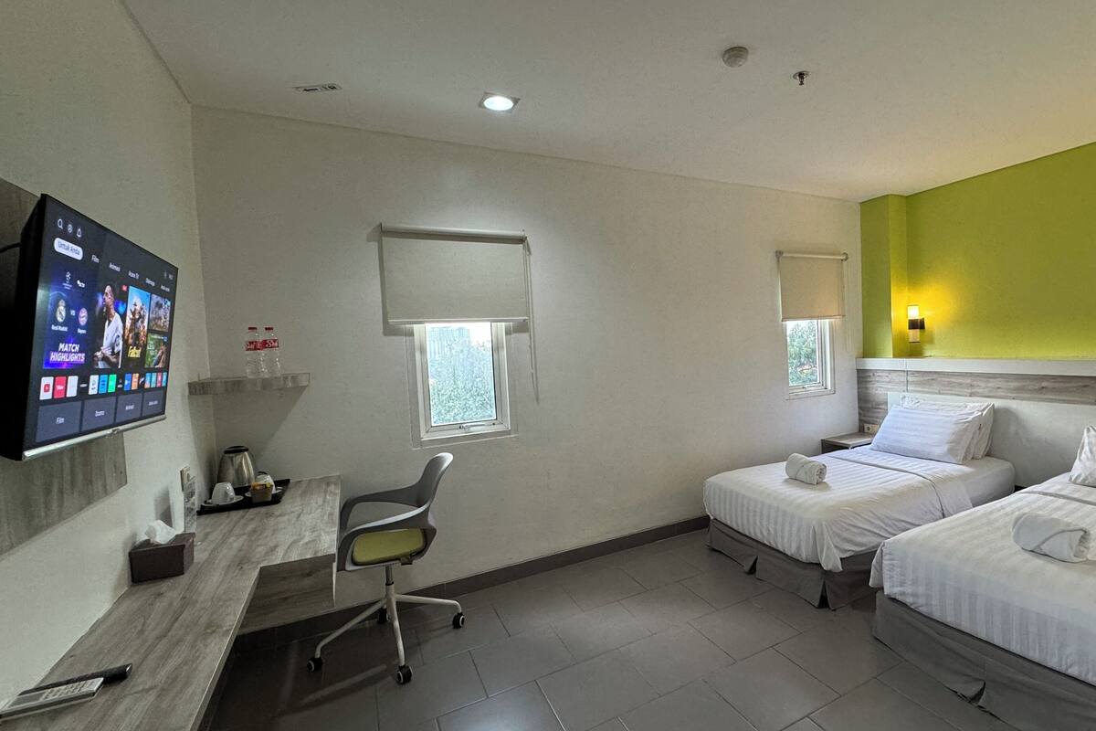 A hotel room with two beds, a desk with a chair, a TV mounted on the wall, and a window with a view of the city.