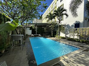 A swimming pool with a tiled deck, surrounded by greenery and a building with multiple windows. - IZI Hotels (Bogor)