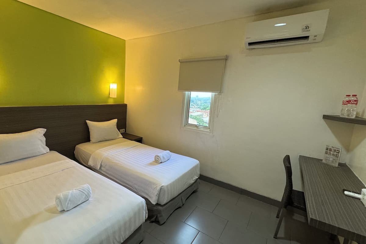 A hotel room with two beds, a small table, a window with a view, an air conditioning unit, and a wall-mounted shelf with a bottle.