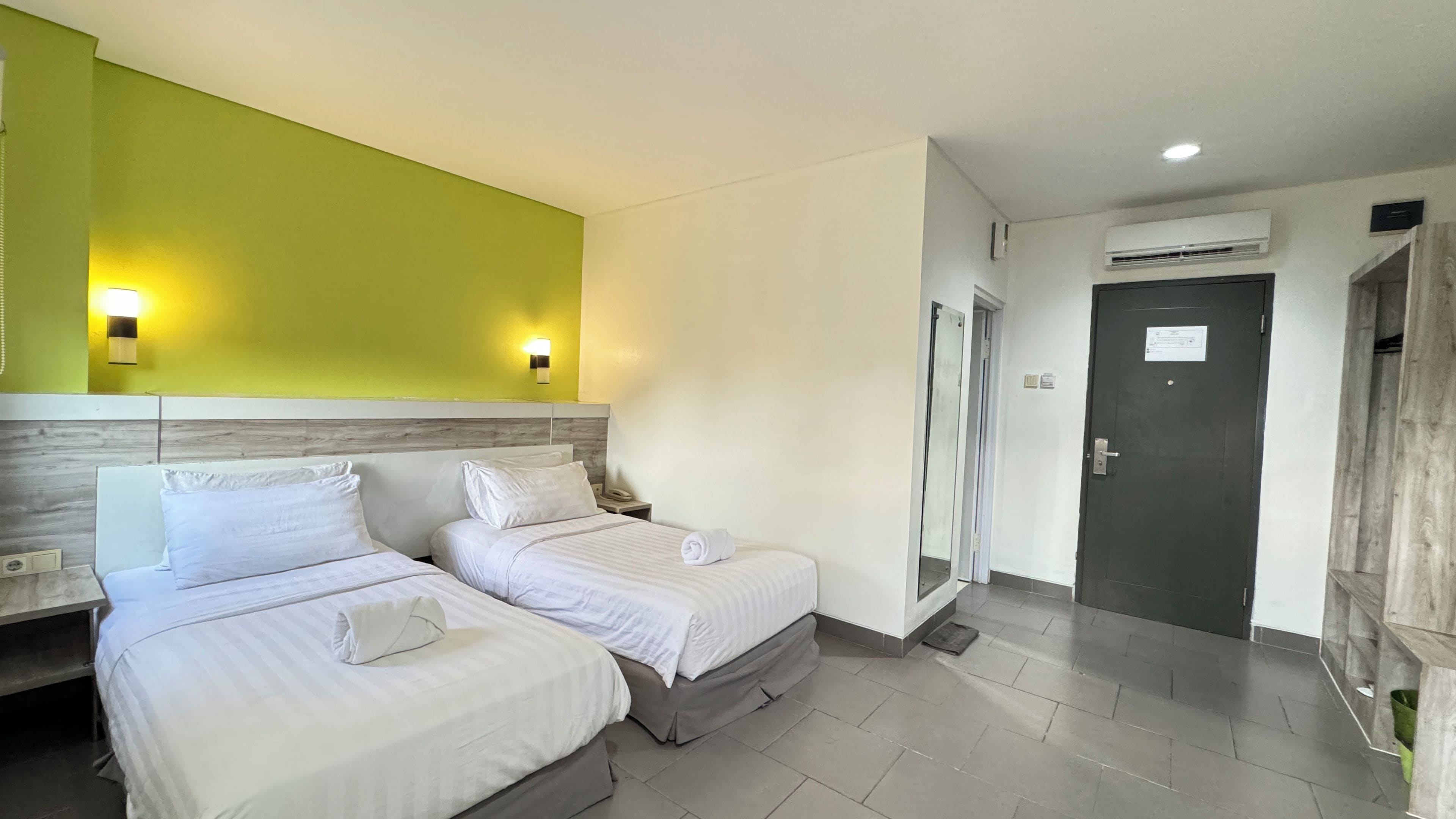 A hotel room with two beds, a green accent wall, and a door leading to a bathroom.