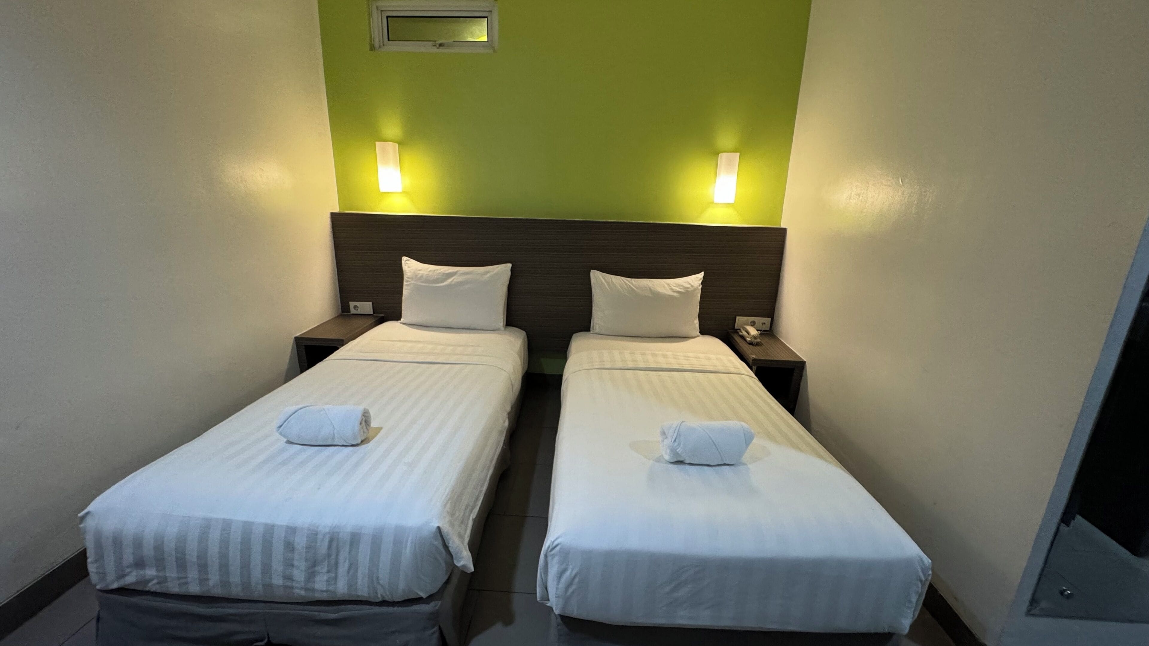 Une chambre d’hôtel avec deux lits simples, chacun muni d’un oreiller et d’une serviette pliée. Il y a des tables de chevet avec des lampes et une petite horloge. Les murs sont peints en vert, et il y a une fenêtre donnant sur l’extérieur.