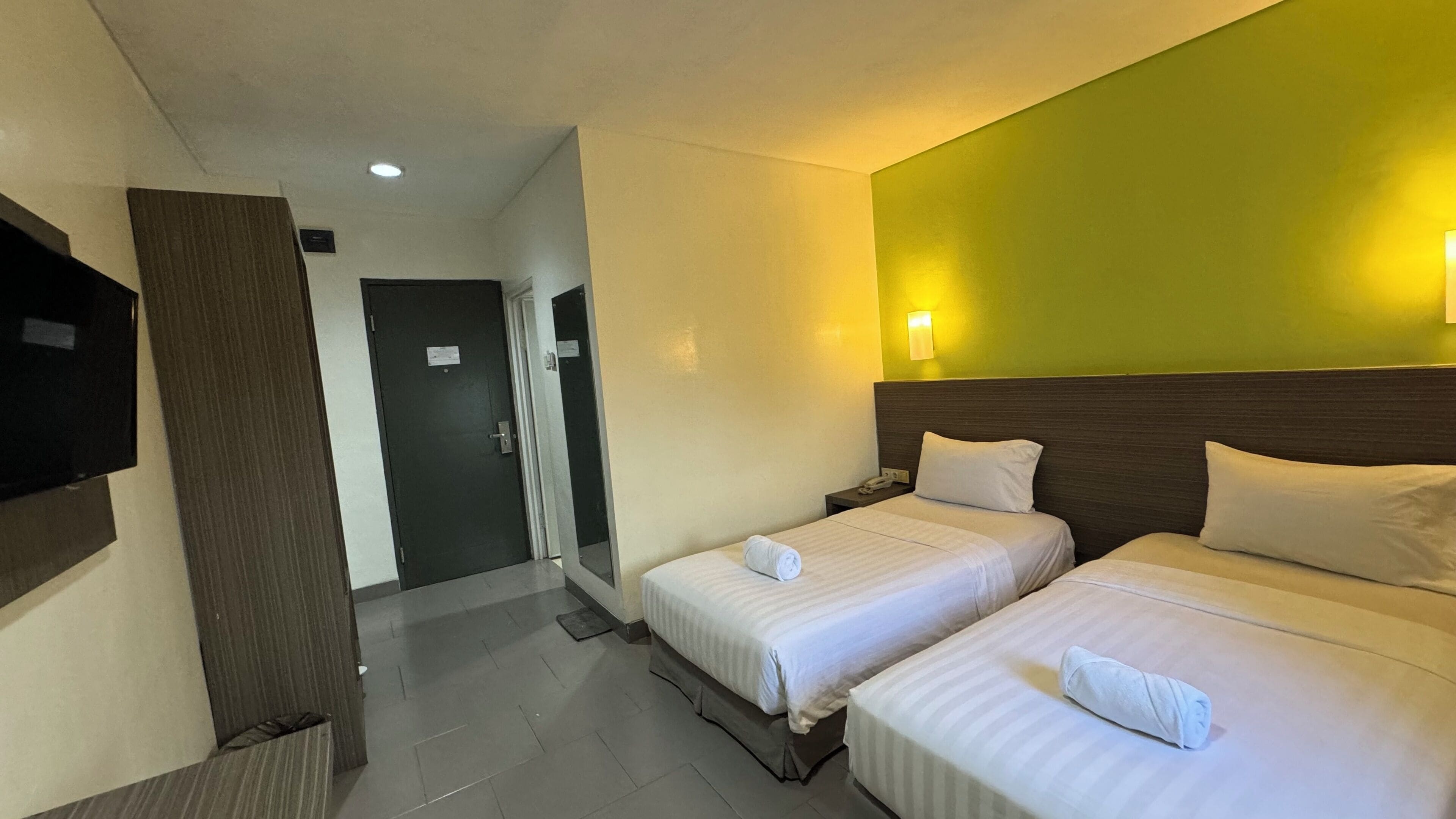 A hotel room with two beds, a television, a wall-mounted light, and a door leading to a hallway.